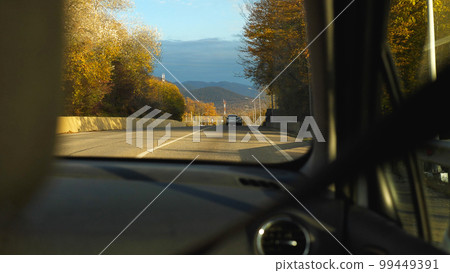 View of the country road from the passenger seat of the car. A beautiful autumn landscape with blue sky, mountains and yellow trees through the windscreen. Road Trip. 99449391
