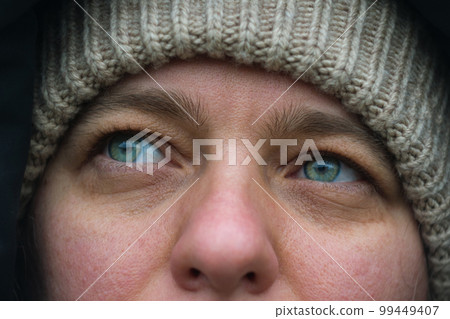 Close-up of the face of a Caucasian girl with beautiful green-blue eyes. A woman in the street wearing a knitted hat. The woman's gaze is dreamily turned to the side. Close-up of the face of a Caucasian girl with beautiful green-blue eyes. A woman in the street wearing a knitted hat. The woman's gaze is dreamily turned to the side. 99449407