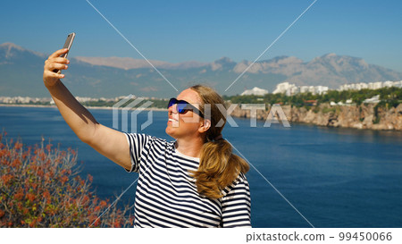 Close-up of a girl taking selfies with her smartphone against a beautiful seascape with mountains. Young woman with sunglasses taking selfies against the sea. Close-up of a girl taking selfies with her smartphone against a beautiful seascape with mountains. Young woman with sunglasses taking selfies against the sea. 99450066