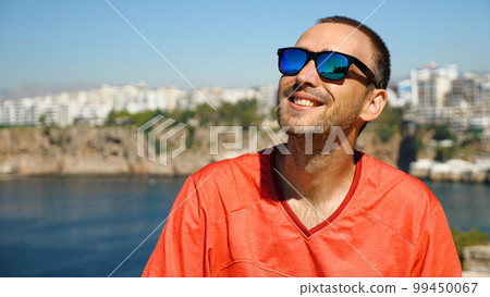 Handsome man with sunglasses in the background - sea and city. Caucasian man in red jersey in a beautiful seascape with mountains in the background. 99450067