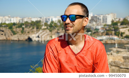 Handsome man with sunglasses smiling against a background of sea and city. Caucasian man in red jersey against a beautiful seascape with mountains. Handsome man with sunglasses smiling against a background of sea and city. Caucasian man in red jersey against a beautiful seascape with mountains. 99450070
