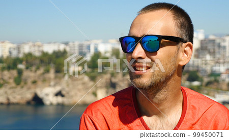 A handsome man with sunglasses smiling against the background of the sea and the city. Caucasian man in red jersey against a beautiful seascape with mountains. A handsome man with sunglasses smiling against the background of the sea and the city. Caucasian man in red jersey against a beautiful seascape with mountains. 99450071