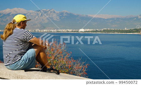 A girl is sitting on a balustrade on the edge of a cliff in front of the mountains, looking out to sea. A lonely girl on holiday enjoying the view of the Mediterranean Sea. A girl is sitting on a balustrade on the edge of a cliff in front of the mountains, looking out to sea. A lonely girl on holiday enjoying the view of the Mediterranean Sea. 99450072