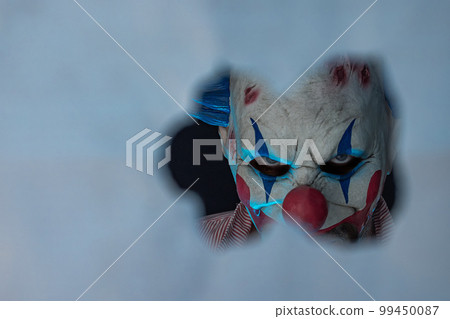 Close-up of a creepy clown looking through a hole. The face of a man wearing a creepy clown mask, looking at his victim. Blue lighting of the crazy clown. 99450087