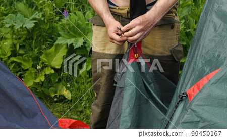 Close-up of a man's hands tying a knot in the tent. In his hand is the tourist aluminium peg. Pitching the tent. Hiking. Sports rest. Travel and tourism. Close-up of a man's hands tying a knot in the tent. In his hand is the tourist aluminium peg. Pitching the tent. Hiking. Sports rest. Travel and tourism. 99450167