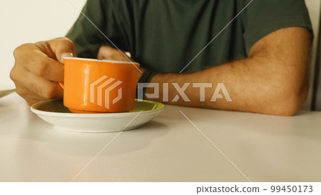 A man's hand is holding an orange mug of hot coffee over a saucer. Steam comes out of the mug. Close up. A man is drinking coffee in a cafe. 99450173