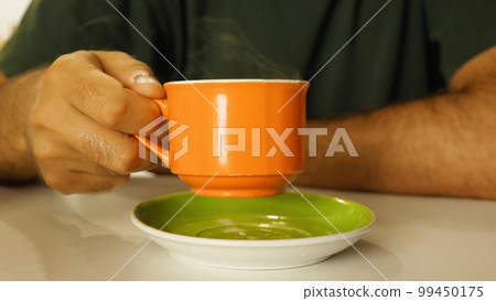 A man's hand is holding an orange mug of hot coffee, with steam coming out of it, over a green saucer. Close up. Man drinking coffee in a cafe. 99450175