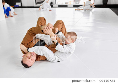 Dynamic portrait of man, professional judo, jiu-jitsu coach training with little boy, child in white kimono. Teaching kids endurance and strength 99450783