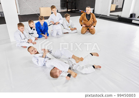 Boys, children in kimonos sitting in circle and watching training judo, jiu-jitsu activity. Learning sport tricks with professional coach Boys, children in kimonos sitting in circle and watching training judo, jiu-jitsu activity. Learning sport tricks with professional coach 99450803