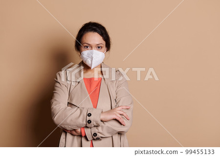 Confident multi-ethnic pretty woman, a businesswoman entrepreneur manager, wearing protective medical mask, standing with her hands folded, looking at camera, over isolated beige background. Ad space 99455133