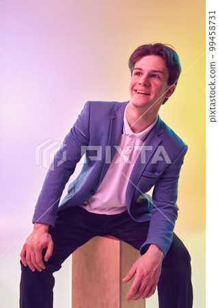 Portrait of happy handsome young man, office clerk, student in jacket and jeans posing over light gradient yellow-purple background. Fashion, art, education, business concept 99458731
