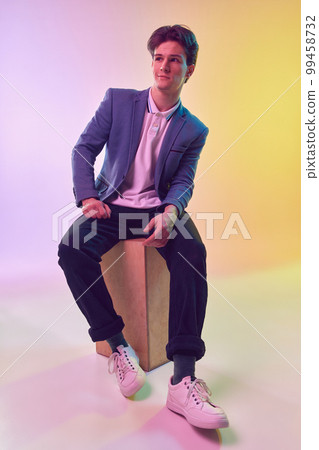 Young handsome man, office clerk, student in jacket and jeans sitting on box over light gradient yellow-purple background. Fashion, art, education, business concept 99458732