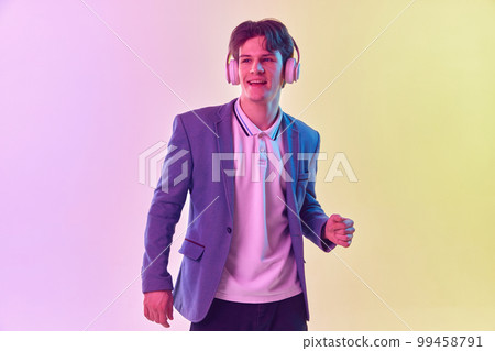 Half-length portrait of young man in headphones dancing isolated on gradient yellow-purple background in neon light. Concept of human emotions, youth culture. 99458791