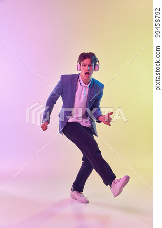 Full-length portrait of young man in headphones dancing isolated on gradient yellow-purple background in neon light. Concept of human emotions, youth culture. 99458792