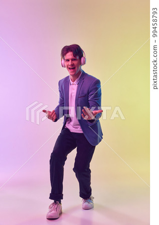 Full-length portrait of young man in headphones dancing isolated on gradient yellow-purple background in neon light. Concept of human emotions, youth culture. 99458793