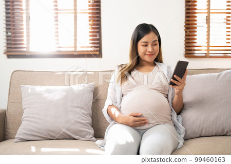 Happy Asian pregnant woman using her phone while relaxing on sofa in her living room 99460163