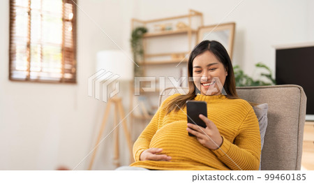 Happy Asian pregnant woman using her phone while relaxing on sofa in her living room Happy Asian pregnant woman using her phone while relaxing on sofa in her living room 99460185