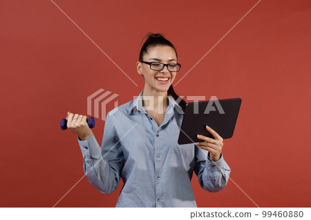 Successful young modern businesswoman in blue shirt warming up with dumbbell at office while working on tablet pad. Multitasking and time management concept. 99460880