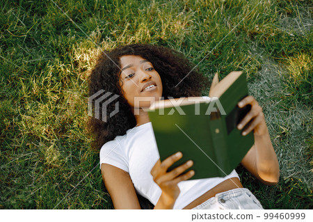 African girl reading a book while lying on a grass. Focus on a girl's face. Girl wearing a white clothes. 99460999