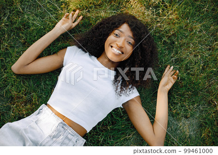 African girl looking at the camera while lying on a grass. Girl is posing for a photo. Girl wearing a white clothes. 99461000