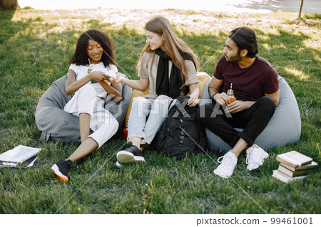 Friends hanging in the green park, relaxing. Boy and girls holding a bottles of juice. African girl wearing white clothes, boy wearing brown t-shirt and black jeans. 99461001