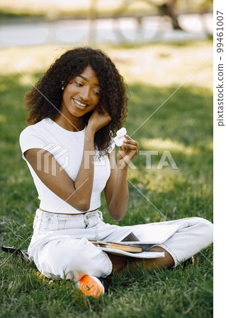 African girl holding a earpods while sitting on a grass. Girl is using a white tablet. Girl wearing a white clothes. African girl holding a earpods while sitting on a grass. Girl is using a white tablet. Girl wearing a white clothes. 99461007