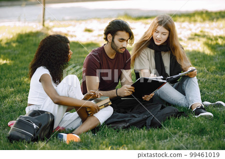 Group of international students sitting on a grass together in park at university. African and caucasian girls and indian boy talking outdoors. Three friends disscussing about education. Group of international students sitting on a grass together in park at university. African and caucasian girls and indian boy talking outdoors. Three friends disscussing about education. 99461019