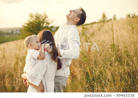 Photo of a young family walking at the field on a sunny day. Brunette mother, father and their little blonde son posing for a photo. Mother holding her son on a hands. Photo of a young family walking at the field on a sunny day. Brunette mother, father and their little blonde son posing for a photo. Mother holding her son on a hands. 99461036