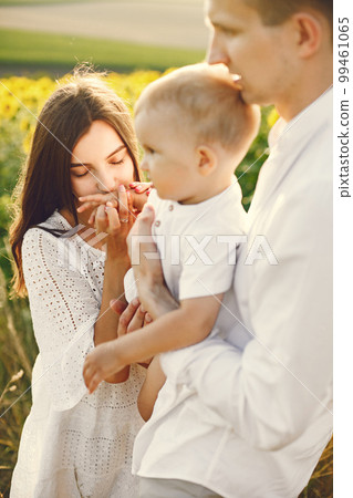 Photo of a young family at the sunflowers field on a sunny day. Brunette mother, father and their little blonde son posing for a photo. Man and woman hugging their son. 99461065