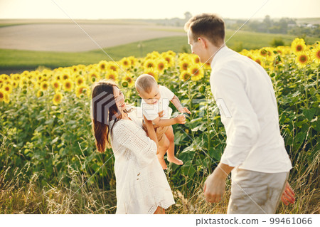 Photo of a young family at the sunflowers field on a sunny day. Brunette mother, father and their little blonde son posing for a photo. Man and woman playing with their son. 99461066