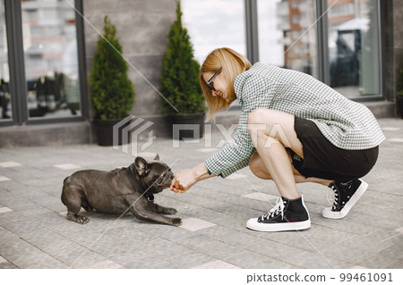 Young woman sitting on a pavement outdoors with black French bulldog. Girl wearing black sunglasses, shorts and grey jacket. Woman playing with a dog. 99461091