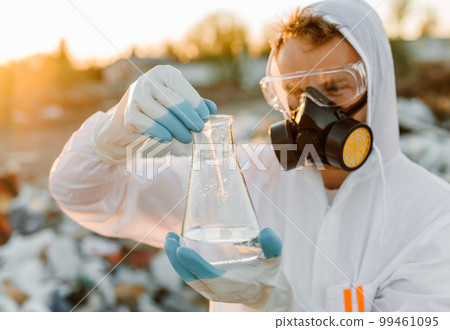 Male ecologist in radiation suit, gas mask. Holding test tube with liquid while studying trash dump. 99461095