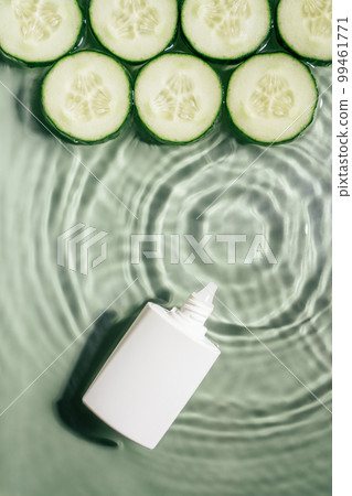 White beauty product cream vial tube with row of cucumber slices on surface of rippled transparent green water. Vertical 99461771