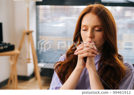 Close up portrait of sorrow young woman sincerely praying with eyes closed holding folded hands in front of face at home by window. Worried female put hands in prayer praying excitedly with hope. Close up portrait of sorrow young woman sincerely praying with eyes closed holding folded hands in front of face at home by window. Worried female put hands in prayer praying excitedly with hope. 99461912