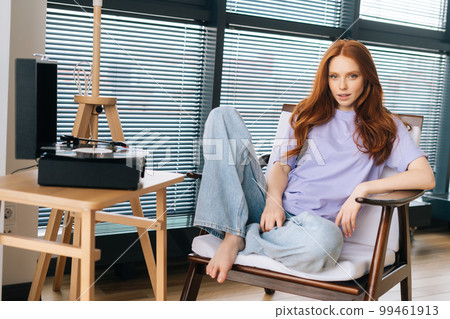 Portrait of charming young woman enjoying listening music on old gramophone vinyl record player sitting on chair looking at camera. Female enjoying home entertainment, using retro media technology. 99461913