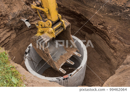 Excavator with a bucket, lowering into the pit on steel cables concrete sewer ring. Construction or repair of sewer home 99462064