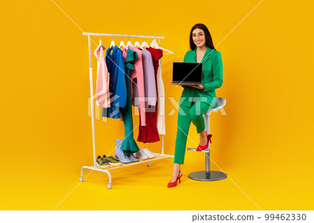 Online shopping. Happy female shopaholic showing laptop screen for retail website ad, sitting near clothing rail 99462330