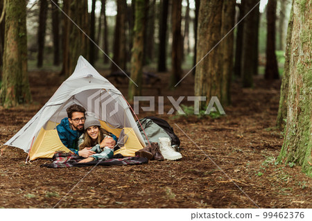 Smiling millennial caucasian guy and woman in jackets rest in cold forest, enjoy adventure lies in tent Smiling millennial caucasian guy and woman in jackets rest in cold forest, enjoy adventure lies in tent 99462376
