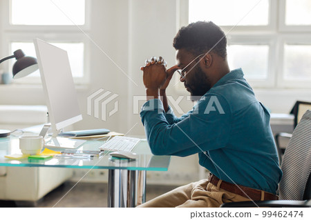 Thoughtful black male entrepreneur in glasses sitting at desk in front of computer, working online in office, side view Thoughtful black male entrepreneur in glasses sitting at desk in front of computer, working online in office, side view 99462474