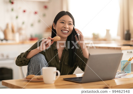 Cheerful asian woman using laptop at kitchen, talking on phone 99462548