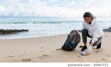 Young female volunteer picking bottles into plastic bag black for cleaning beach in morning time, panorama, free space Young female volunteer picking bottles into plastic bag black for cleaning beach in morning time, panorama, free space 99462552