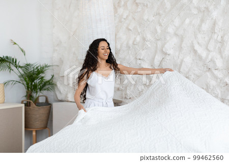 Cheerful millennial caucasian lady brunette in pajamas make bed, enjoy good morning, hold blanket Cheerful millennial caucasian lady brunette in pajamas make bed, enjoy good morning, hold blanket 99462560