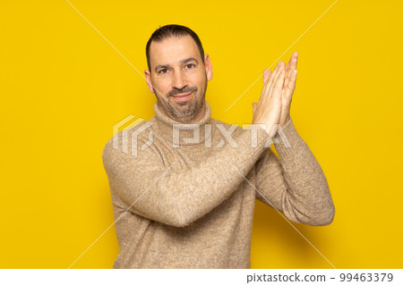 Bearded hispanic man in his 40s wearing a beige turtleneck clapping his hands in satisfaction with his hands to the side, isolated over yellow background. Bearded hispanic man in his 40s wearing a beige turtleneck clapping his hands in satisfaction with his hands to the side, isolated over yellow background. 99463379