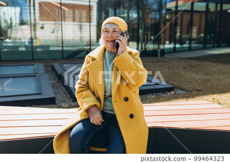 Old woman talking of the her phone. Senior blonde woman has a happy conversation at cellphone. Smiling woman using phone sitting on the bench on sity street. Old woman talking of the her phone. Senior blonde woman has a happy conversation at cellphone. Smiling woman using phone sitting on the bench on sity street. 99464323