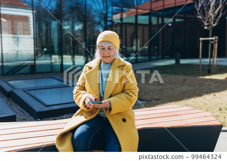 Beautiful senior woman in casual clothing using her smart phone while sitting on the bench on sity street. 99464324