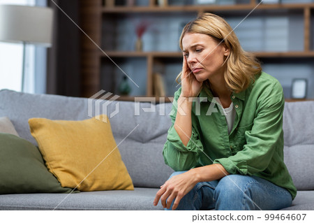 Mature woman alone at home with boss and depressed, housewife sitting upset on sofa holding head with hands in living room. 99464607