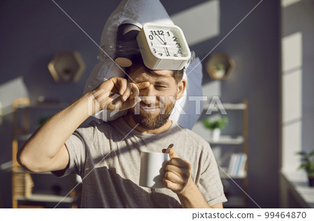 Man with a pillow and an alarm clock drinking morning coffee and rubbing his eyes 99464870