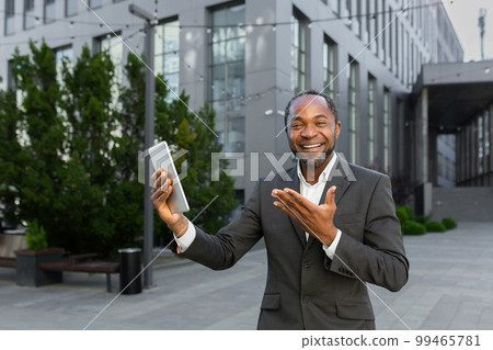 An African-American businessman is standing outside in a suit, holding a tablet and talking on a video call. He looks at the camera, points to the gadget, smiles. An African-American businessman is standing outside in a suit, holding a tablet and talking on a video call. He looks at the camera, points to the gadget, smiles. 99465781
