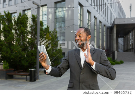 An African-American businessman is standing outside in a suit, holding a tablet and talking on a video call. An African-American businessman is standing outside in a suit, holding a tablet and talking on a video call. 99465782