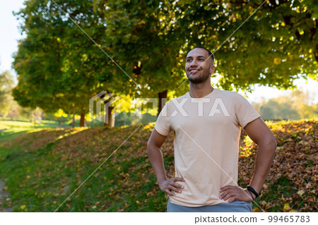 Portrait of a young African-American athlete standing in a park in a sports uniform. Runner, athlete, morning run. The smiling man looks to the side, holds his hands on his waist. 99465783